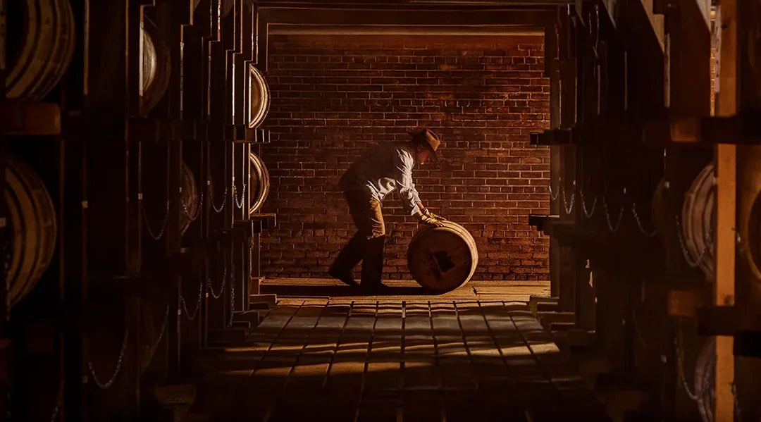 A worker rolling a barrel down a dim rickhouse aisle