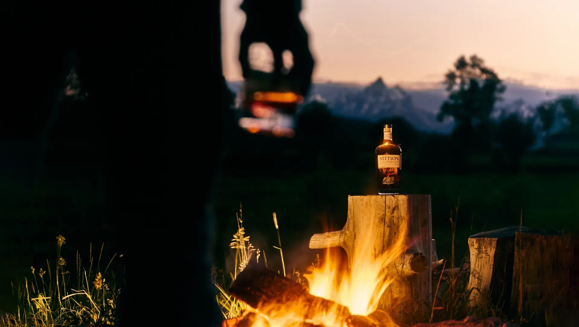A bottle of Stetson Bourbon on a stump beside a campfire at dusk, mountains behind